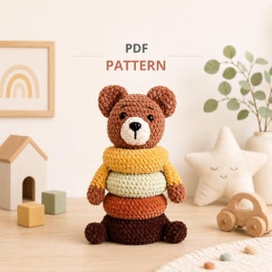 May include: A crochet bear stacking toy made of soft brown, yellow, and cream yarn. The bear has a round head with black eyes and a white muzzle, sitting atop three textured rings in yellow, light green, and orange. Text reads PDF PATTERN.