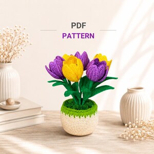 Puede incluir: Un arreglo de tulipanes de ganchillo con flores moradas y amarillas, hojas verdes y una maceta color crema y verde. El texto PDF PATTERN aparece sobre las flores. El arreglo está rodeado de jarrones blancos y flores secas.