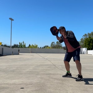 May include: A person wearing a black jacket, red shirt, and blue shorts is playing pickleball on a rooftop parking lot. They are holding a black pickleball paddle and a yellow ball is flying through the air.