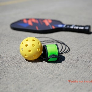 May include: A yellow pickleball with holes and a green and black paddle grip on a gray concrete surface. *Paddle not included*