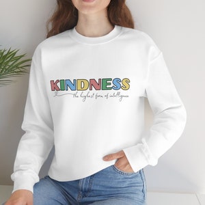 Puede incluir: Sudadera blanca con la palabra "KINDNESS" en letras de colores y el texto "la forma más alta de inteligencia" en letra cursiva.