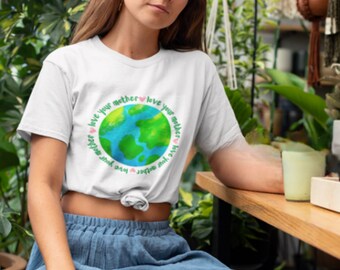 May include: A white t-shirt with a green and blue earth graphic and the text "Love your mother love your mother love your mother love your mother" in a circle around the earth. The shirt is tied at the waist and the person wearing it is sitting on a wooden bench.