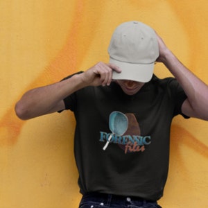 May include: A person wearing a black t-shirt with a blue and orange graphic of a tennis racket and the text "Forensic Files". The person is wearing a beige baseball cap.