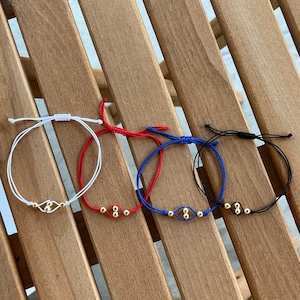 Peut inclure: Quatre bracelets réglables en blanc, rouge, bleu et noir. Chaque bracelet est orné d'un charm doré avec trois petites perles et une fermeture coulissante. Les bracelets sont présentés sur une surface en bois.