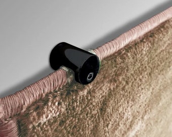 May include: A black rug clip is attached to the edge of a beige rug. The clip is made of shiny black material and is designed to hold the rug in place. The rug has a textured surface and a pink border.