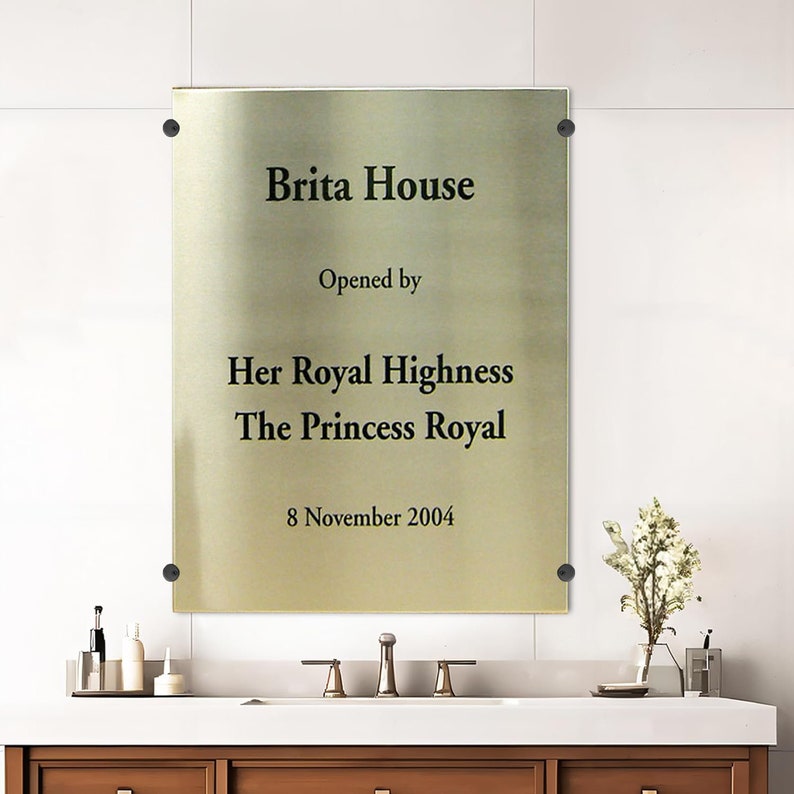 May include: A rectangular gold-colored plaque with the text "Brita House" and "Opened by Her Royal Highness The Princess Royal 8 November 2004". The plaque is mounted on a white wall with black fasteners.