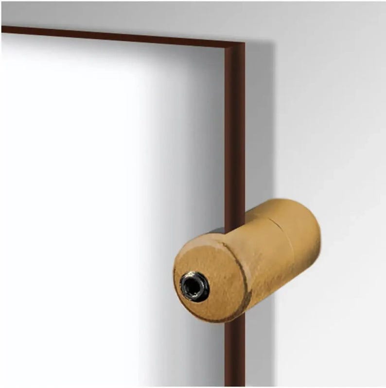 May include: A light brown, cylindrical object with a black center, possibly a door knob or furniture hardware, is attached to a brown rectangular frame. The frame is set against a white wall.