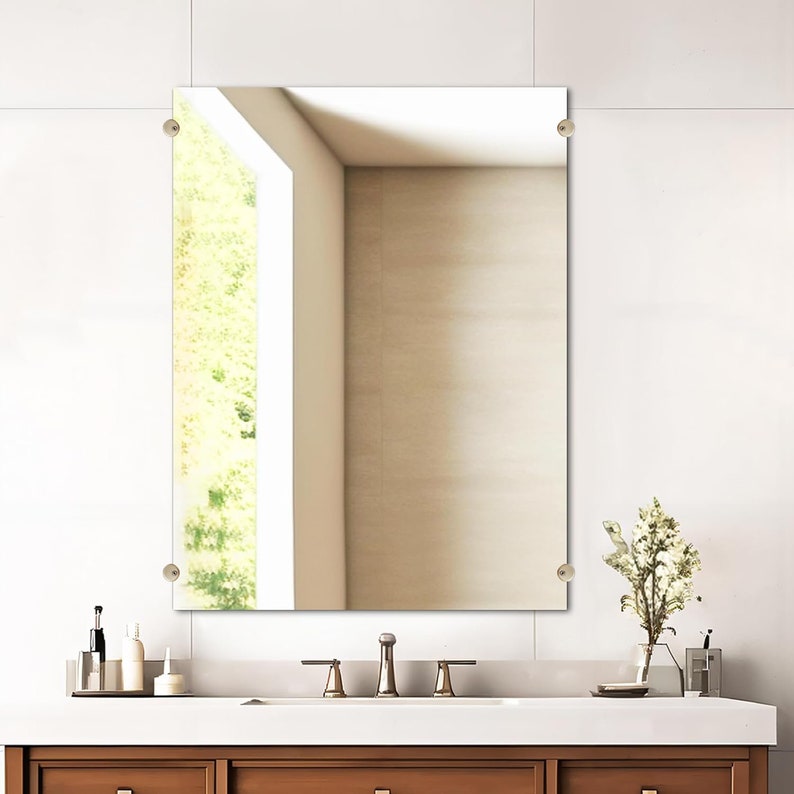 May include: A rectangular frameless bathroom mirror with rounded corner hardware. The mirror is mounted on a white wall above a brown vanity with a white countertop. A window is visible in the reflection.