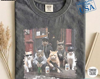 May include: Pepper-colored Comfort Colors t-shirt featuring a graphic of six cats dressed in urban attire. The cats are depicted with sunglasses, hats, and holding items like a beer bottle and a toy gun. The text "up 2-3 size for Oversized" is at the bottom.