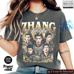 May include: Dark gray t-shirt featuring the name "ZHANG Linghe" in gold lettering, with a graphic of five men. The shirt has the text "COMFORT COLORS" and "Pepper color" printed on it. The shirt is oversized and the text "up 2-3 size for Oversized" is printed on the bottom.