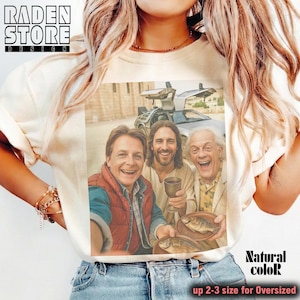May include: Cream-colored t-shirt featuring a selfie of three smiling men, one holding a chalice and another holding a plate of fish, with a DeLorean car in the background. The shirt has the text "Natural color" and "up 2-3 size for Oversized".