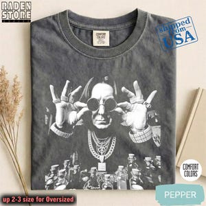 May include: Pepper-colored Comfort Colors t-shirt featuring a black and white graphic of a person wearing sunglasses and jewelry. The text "up 2-3 size for Oversized" is at the bottom. The shirt has a crew neck.