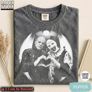 May include: A charcoal gray crew neck t-shirt featuring a black and white graphic of Pinhead and Art the Clown forming a heart shape with their hands. The shirt has a Comfort Colors label at the inner neckline and a vintage-style washed finish.