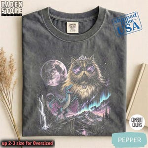 Könnte beinhalten: Ein anthrazitfarbenes T-Shirt mit einer Grafik einer flauschigen Katze, die eine Krone und eine runde Sonnenbrille trägt, über einem blauen Dinosaurier mit violetten Flügeln. Die Szene zeigt einen Vollmond, einen Wasserfall, Berge und Polarlichter.
