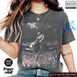 May include: Dark gray t-shirt featuring a graphic of a basketball player in action, with a basketball hoop and a crowd in the background. The shirt has the text "COMFORT COLORS" and "Pepper color". The shirt is oversized.