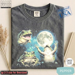 Può includere: T-shirt Comfort Colors grigio scuro con un design stravagante di tre rane verdi, una luna piena e uno sfondo stellato. La maglietta ha il testo "PEPPER" in basso.