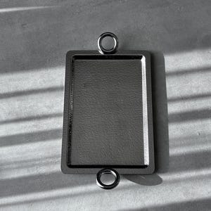 Könnte beinhalten: Ein schwarzes Tablett aus Metall mit zwei runden Griffen. Das Tablett hat eine strukturierte Oberfläche und eine rechteckige Form.