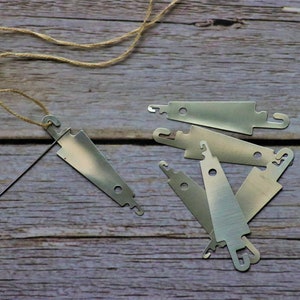 Könnte beinhalten: Eine Sammlung silberfarbener Metallwebwerkzeuge, darunter eine Nadel mit Faden. Die Werkzeuge liegen auf einer Holzoberfläche, was auf ein Bastel- oder DIY-Projekt hindeutet. Die Werkzeuge haben eine einzigartige Form mit Haken und Loch.