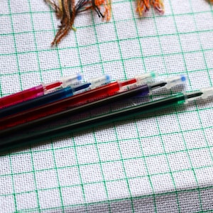 May include: A close-up of several colorful embroidery pens with clear caps, arranged diagonally across a white fabric with a green grid pattern. The pens are red, blue, green, and purple. Embroidery supplies.