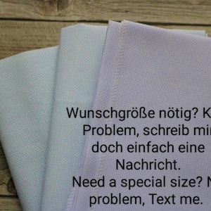 Op de afbeelding: Drie gevouwen stoffen stukken in blauwe en lavendel tinten zijn op een houten oppervlak gestapeld. De tekst op de stof luidt: "Wunschgröße nötig? Kein Problem, schreib mir doch einfach eine Nachricht."