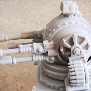 Könnte beinhalten: Eine hellgraue Harzminiatur eines kugelförmigen Geschützturmsystems. Das Modell verfügt über drei hervorstehende Läufe, einen kreisförmigen seitlichen Zahnradmechanismus und einen Munitionsgurt, der in die Basis führt, montiert auf einer quadratischen Plattform.