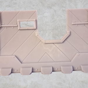 Può includere: Un modello di edificio in plastica marrone chiaro con una finestra e una superficie testurizzata. Il modello presenta una serie di piccole sporgenze arrotondate lungo il bordo inferiore.