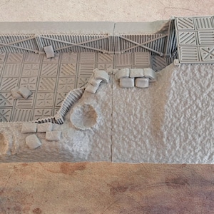 Może przedstawiać: Szczegółowa, jasnoszara miniatura dioramy przedstawiająca fragment fortecy lub budynku. Model posiada teksturowane ściany, wzorzystą podłogę oraz detale architektoniczne, takie jak rampa i belki podporowe. Ogólny projekt sugeruje motyw fantasy lub historyczny.