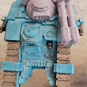 May include: A detailed, top-down view of a model tank. The tank is primarily light blue with a textured surface and features a large turret and multiple gun barrels. The treads are a dark, metallic color.