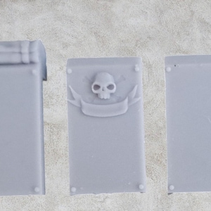 Può includere: Tre targhe decorative rettangolari grigie. La prima ha una parte superiore arrotolata, la seconda presenta un design con teschio e banner, e la terza è semplice. Ogni targa ha piccoli dettagli circolari agli angoli.