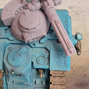 May include: A detailed, light blue model of a military vehicle with a large gun turret and multiple barrels. The model has a textured surface and is painted in shades of blue and gray. The gun is a light gray color.