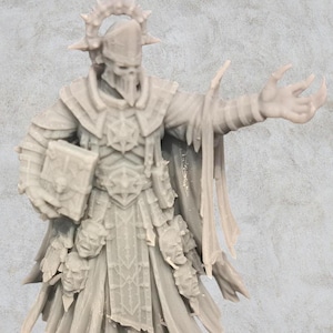 Puede incluir: Una miniatura de resina gris claro sin pintar de una figura esquelética con armadura ornamentada, un tocado alto con un halo de púas y una túnica larga adornada con múltiples caras pequeñas esculpidas. La figura sostiene un libro grueso.