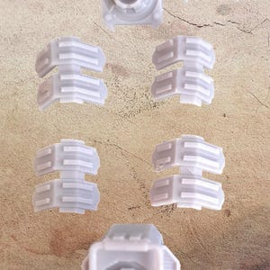 Pode incluir: Uma variedade de componentes de plástico cinza claro, incluindo várias peças segmentadas, uma peça circular com um design central e uma peça curva semicircular. As peças estão dispostas sobre uma superfície texturizada de tom neutro.