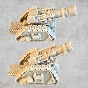 Puede incluir: Un par de cañones en miniatura de plástico gris sobre orugas. Los cañones apuntan hacia arriba y tienen un diseño detallado.