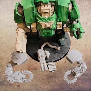 Puede incluir: Modelo de máquina de guerra en miniatura verde y blanquecino parcialmente ensamblado. El modelo presenta un torso grande, una pierna y dos brazos con accesorios de sierra circular. Componentes adicionales están dispersos alrededor de la base.