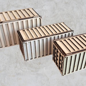 Può includere: Tre scatole di legno a forma di container, di colore marrone chiaro con bordi marrone scuro. Le scatole sono rettangolari con doghe verticali sui lati e un coperchio a doghe.