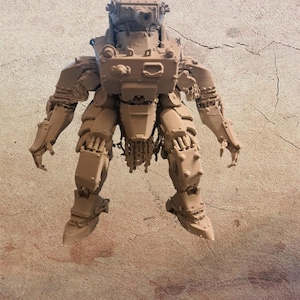 Puede incluir: Una figura de robot mecánico impresa en 3D de color tostado, con un torso en forma de torreta de tanque con un cañón montado y una pequeña figura de soldado en la parte superior. La máquina bípeda tiene extremidades articuladas y manos en garra.