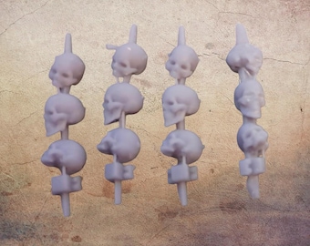May include: Four light purple skull-shaped decorative skewers. Each skewer has four skulls stacked on a vertical post with a small base. The skulls have detailed features and are evenly spaced. The background is a textured, tan surface.