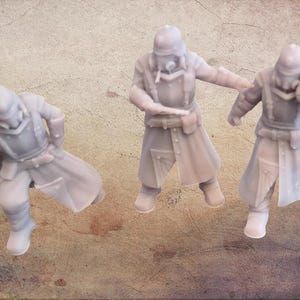 Puede incluir: Tres figuras en miniatura grises de estilo postapocalíptico, cada una con casco, máscara de gas y abrigo largo. Las figuras están en poses dinámicas, sugiriendo acción o movimiento. El fondo es de color neutro texturizado.