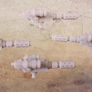 Puede incluir: Cuatro pistolas de juguete en miniatura de color gris claro están dispuestas sobre una superficie texturizada y de color beige. Cada pistola tiene un diseño detallado con componentes cilíndricos y rectangulares, y un pequeño soporte. El tema general es el armamento en miniatura.