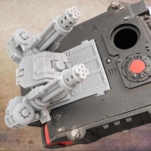 Puede incluir: Una torreta de tanque en miniatura de plástico gris con cañones de múltiples tubos montada sobre un chasis de vehículo blindado negro. El conjunto de la torreta incluye componentes mecánicos detallados y una escotilla roja circular.