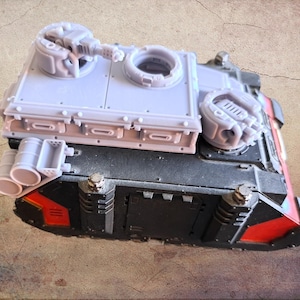 Pode incluir: Modelo detalhado de um tanque militar, com uma estrutura superior cinza claro e um corpo inferior preto com detalhes vermelhos. O tanque tem múltiplas torres, canos e tubos de escape, mostrando elementos de design intrincados.