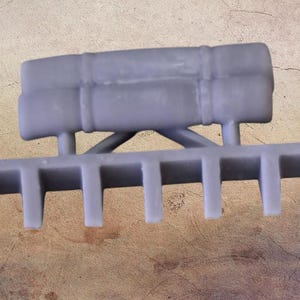 May include: A grey, 3D-printed resin miniature component featuring a horizontal bar with six downward-pointing rectangular teeth. Above the teeth, a cylindrical, log-like structure is attached by two vertical supports and a central cross-brace.