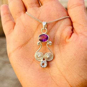 May include: A silver pendant necklace with a purple amethyst gemstone and a clear white topaz gemstone. The pendant features a delicate, swirling design.