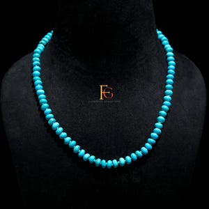 May include: A turquoise bead necklace displayed on a black mannequin. The necklace is composed of small, round, light blue beads. The beads are strung together to form a complete necklace. The background is black.