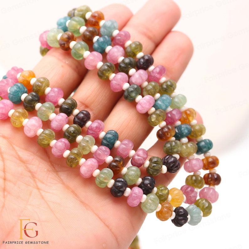 May include: A strand of multi-coloured watermelon tourmaline beads. The beads are round and have a carved, textured surface. The beads are strung on a white thread.