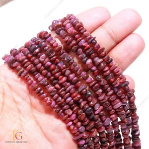 May include: A strand of red ruby gemstone beads, each bead is a small, irregular chip. The beads are strung on a clear thread and are displayed on a hand.