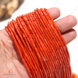 Op de afbeelding: Een streng helder oranje koraal kralen, elke kraal heeft een rechthoekige buisvorm. De kralen zijn aan elkaar geregen en zijn ongeveer 10 mm lang.