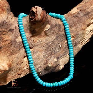 Op de afbeelding: Een turquoise kralenketting afgebeeld op een stuk drijfhout. De ketting heeft kleine, ronde, lichtblauwe kralen die aan elkaar zijn geregen. Het hout heeft een natuurlijk, getextureerd uiterlijk met verschillende tinten bruin en beige.