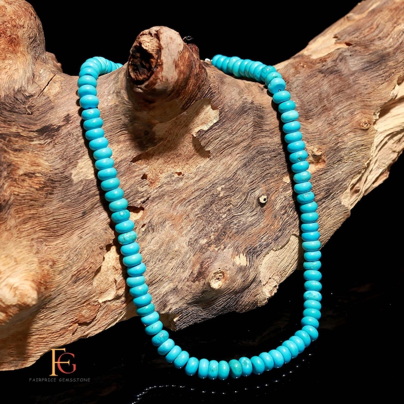 Op de afbeelding: Een turquoise kralenketting afgebeeld op een stuk drijfhout. De ketting heeft kleine, ronde, lichtblauwe kralen die aan elkaar zijn geregen. Het hout heeft een natuurlijk, getextureerd uiterlijk met verschillende tinten bruin en beige.