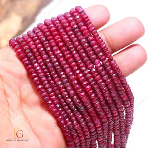 May include: A strand of red ruby beads, faceted and round, strung on a thread. The beads are approximately 4mm in diameter and are arranged in a straight line. The beads are held in a hand, with the background blurred.